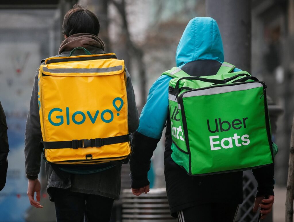 Dois estafetas a caminhar lado a lado, um com uma mochila amarela da Glovo e outro com uma mochila verde da Uber Eats