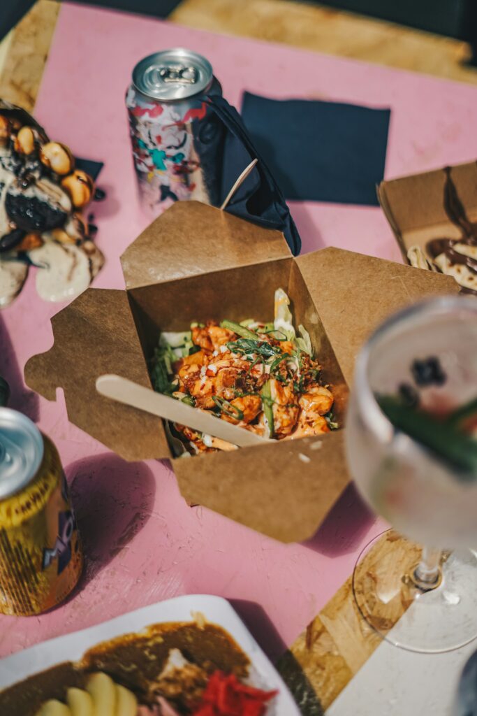 Caixa de cartão aberta com pedaços de frango ou camarão com molho e cebolinho numa mesa com bebidas